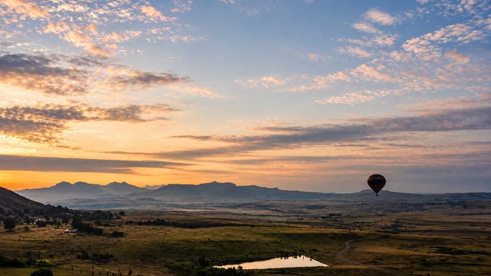 A hot-air balloon drifting over the valley at dawn, with the Maluti mountains beyond